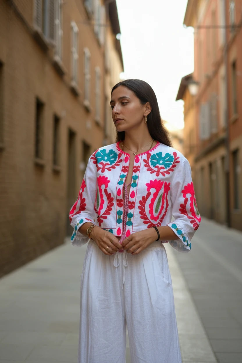 Chaqueta corta blanca Iciar y Candela by Creasur S.L. con bordados florales fucsia y turquesa, mangas acampanadas y cordón ajustable.