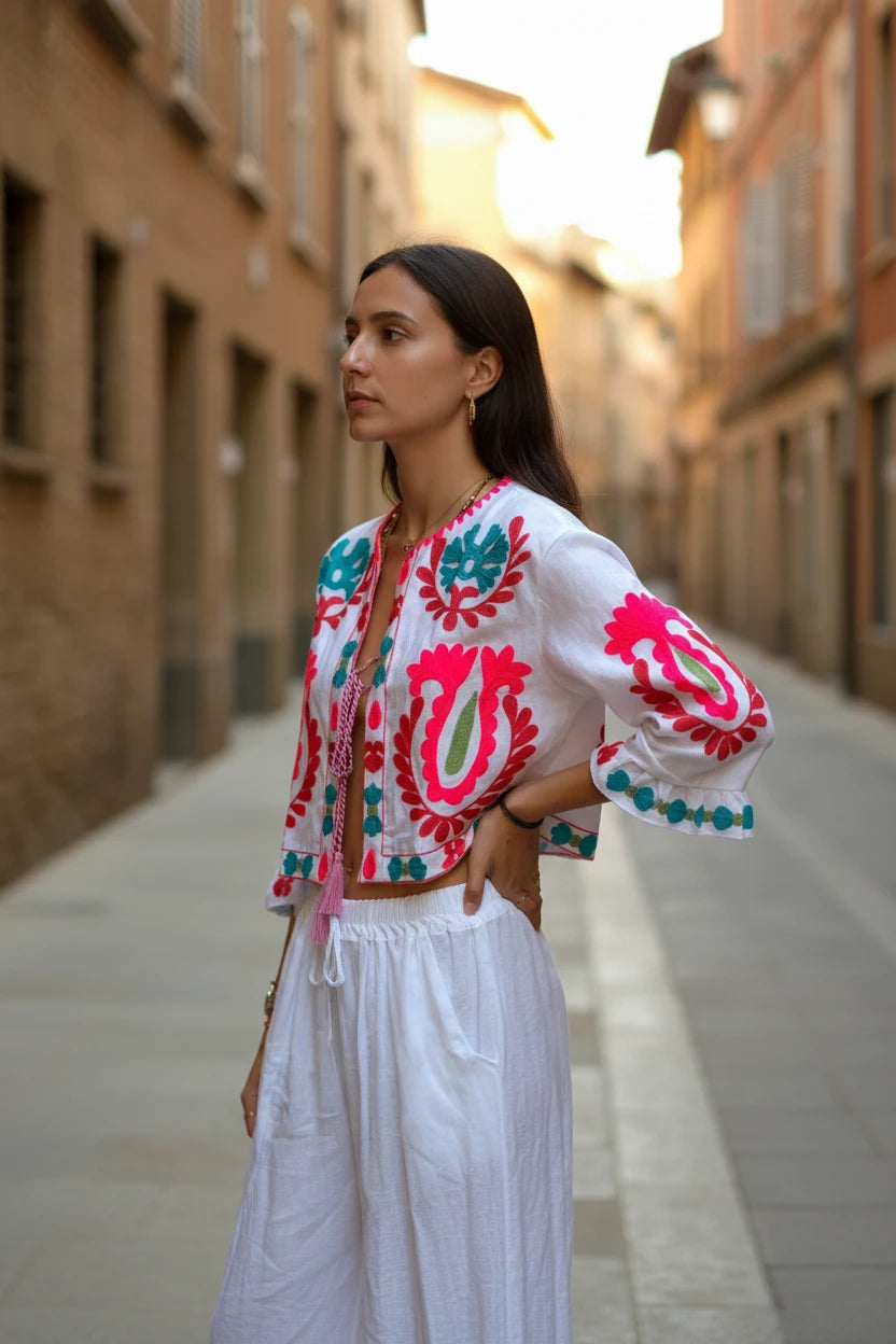 Chaqueta corta blanca Iciar y Candela by Creasur S.L. con bordados florales fucsia y turquesa, mangas acampanadas y cordón ajustable.